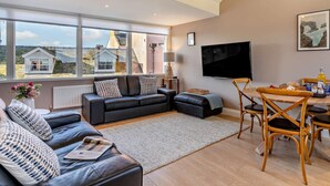Ein modernes Wohnzimmer mit einer Couch, einem Esstisch und Stühlen, einem an der Wand befestigten Fernseher und großen Fenstern mit Blick auf Häuser.