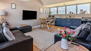 Ein modernes Wohnzimmer mit einer Couch, einem Couchtisch mit Blumenvase, einem Essbereich mit Tisch und Stühlen sowie einer Küche mit blauen Schränken und einem großen Fenster.