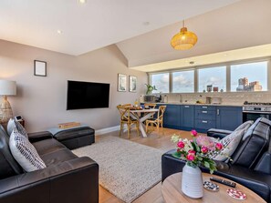 Ein modernes Wohnzimmer mit einer Couch, einem Couchtisch mit Blumenvase, einem Essbereich mit Tisch und Stühlen sowie einer Küche mit blauen Schränken und einem großen Fenster.