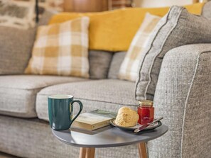 Ein gemütliches Wohnzimmer mit einem grauen Sofa, einem kleinen runden Tisch, einer blauen Tasse, einem Buch, einem Keks und einem Einmachglas Marmelade.