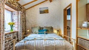 A bedroom with a stone wall, a bed with a metal frame, a window with curtains, and a painting on the wall. - 1 Bed in Bideford (oc-pwork) (Buckland Brewer)
