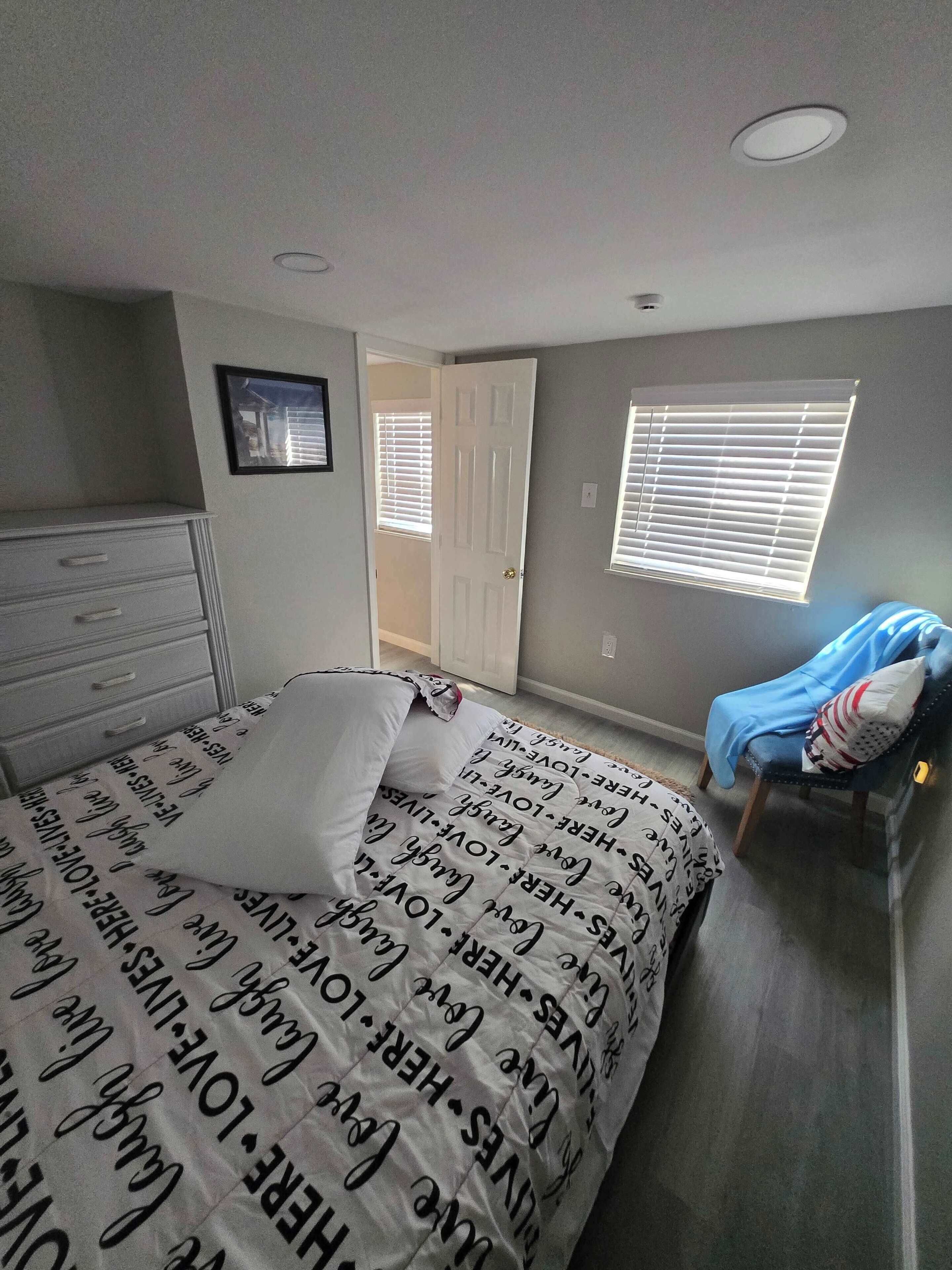 Um quarto com uma cama, cômoda, cadeira, janela com persianas e porta que dá para outro cômodo.