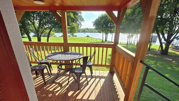 A wooden deck with a table and chairs, overlooking a grassy area with trees and a body of water.