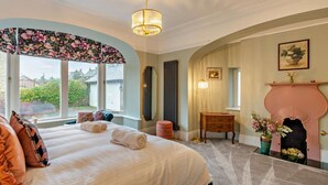 Une chambre avec un grand lit, une fenêtre donnant sur un beau paysage, une cheminée rose et un lustre.