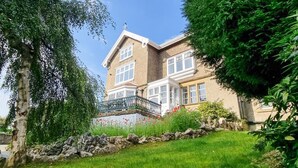 Une maison à deux étages dotée d’un balcon, entourée d’un jardin bien entretenu et d’un mur en pierre.