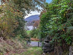 A stone wall with a gate leading to a body of water, surrounded by dense foliage and a hilly landscape in the background. - 2 Bed in Ulverston (oc-91730) (Blawith)