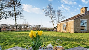 Un espacio al aire libre con césped, una mesa de mimbre dispuesta para té y scones, un jarrón con flores amarillas y un edificio de piedra al fondo.