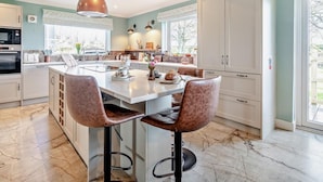Una cocina moderna con una isla central, taburetes de bar, iluminación colgante y vistas al exterior.