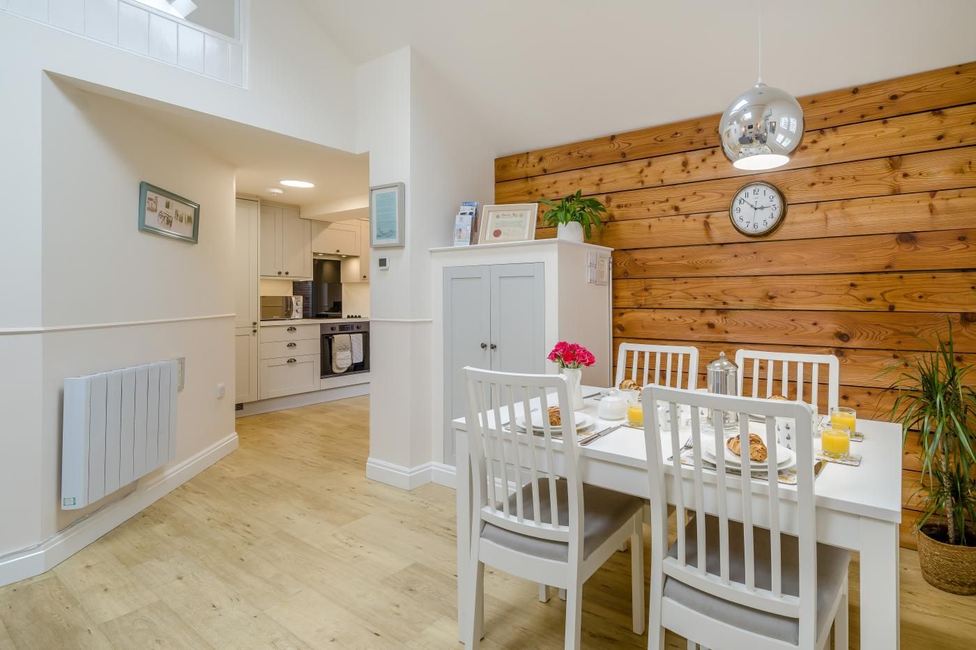 A dining area with white chairs and a table set for breakfast, a wooden wall with a clock, and a kitchen in the background.
