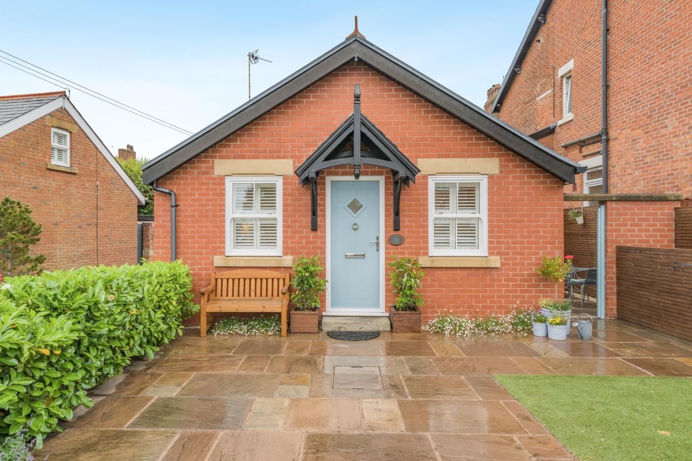 A brick house with a front door, two windows with shutters, a bench, and potted plants.