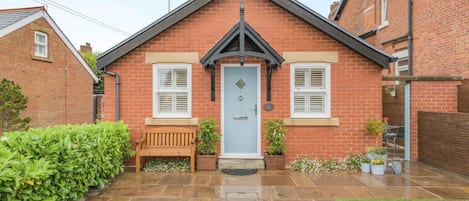 A brick house with a front door, two windows with shutters, a bench, and potted plants.