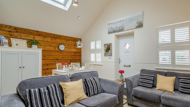 Ein gemütliches Wohnzimmer mit einer Holzwand, einem Esstisch, zwei grauen Sofas mit gestreiften Kissen und einem Bild über der Tür.