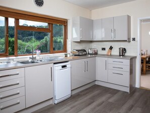 A modern kitchen with a dishwasher, a sink, a window with a view of greenery, and a dining area visible through an open door. - 3 Bed in Brecon (oc-bn419) (Llandeilo'r-Fan)