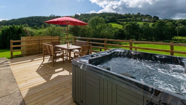 A wooden deck with a hot tub, a table with chairs, and a red umbrella. - 3 Bed in Brecon (oc-bn419) (Llandeilo'r-Fan)
