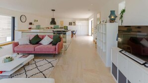 Ein modernes Wohnzimmer mit einem rosafarbenen Sofa, einem Couchtisch und einem Fernseher. Im Hintergrund ist eine Küche mit weißen Schränken und einem Essbereich zu sehen.