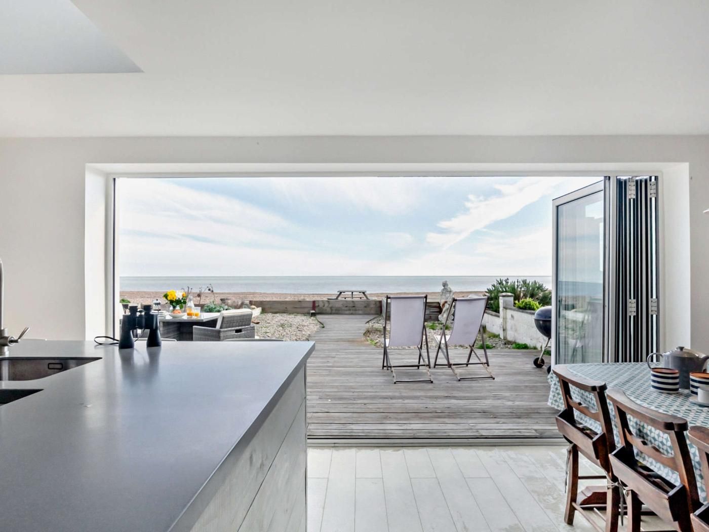 A modern kitchen with a view of the beach, a wooden deck, and outdoor seating.