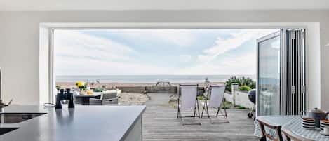 A modern kitchen with a view of the beach, a wooden deck, and outdoor seating.