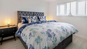 Ein Schlafzimmer mit einem mit Wirbeln besetzten Kopfteil, einem Bett mit floralem Bettzeug, einem Nachttisch mit einer Lampe und einem Fenster mit Jalousien.