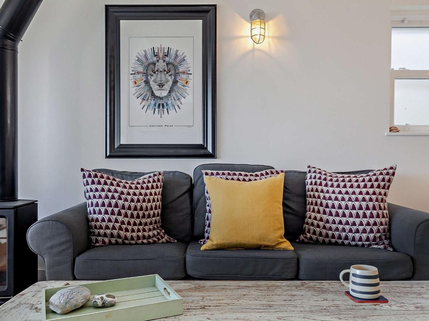 A grey sofa with patterned cushions, a framed artwork, a wall-mounted light, and a rustic coffee table with a mug.