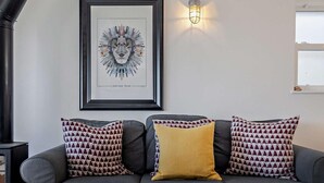 Ein graues Sofa mit gemusterten Kissen, ein gerahmtes Kunstwerk, eine Wandleuchte und ein rustikaler Couchtisch mit einer Tasse.
