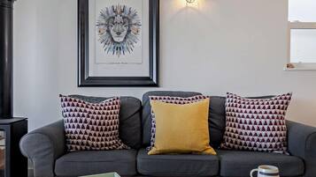 A grey sofa with patterned cushions, a framed artwork, a wall-mounted light, and a rustic coffee table with a mug.