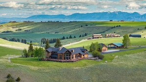 A large house with a deck, surrounded by green fields and mountains in the background. - New Listing | Hot Tub | Secluded Luxury Lodge | Dogs Welcome (Gallatin Gateway)
