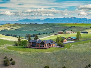 A large house with a deck, surrounded by green fields and mountains in the background. - New Listing | Hot Tub | Secluded Luxury Lodge | Dogs Welcome (Gallatin Gateway)
