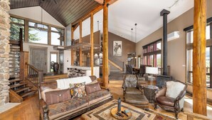 A spacious living area with a stone wall, wooden beams, a fireplace, and a staircase leading to an upper level. - New Listing | Hot Tub | Secluded Luxury Lodge | Dogs Welcome (Gallatin Gateway)