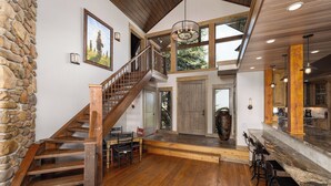 A spacious interior with a wooden staircase, a stone wall, a large wooden door, and a kitchen area with bar stools. - New Listing | Hot Tub | Secluded Luxury Lodge | Dogs Welcome (Gallatin Gateway)