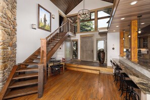A spacious interior with a wooden staircase, a stone wall, a large wooden door, and a kitchen area with bar stools. - New Listing | Hot Tub | Secluded Luxury Lodge | Dogs Welcome (Gallatin Gateway)