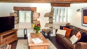 A living room with a television, wooden coffee table, leather sofa, stone walls, and a window with shutters. - 2 Bed in Lancaster (oc-91411) (Clapham)