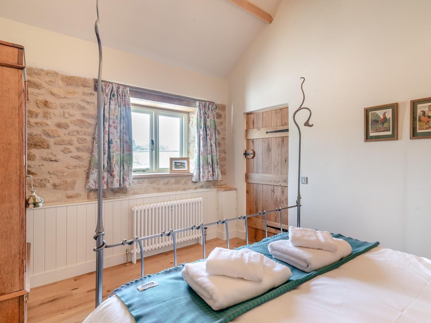 A bedroom with a wooden bed, a radiator, a window with curtains, a wooden wardrobe, and a picture on the wall.
