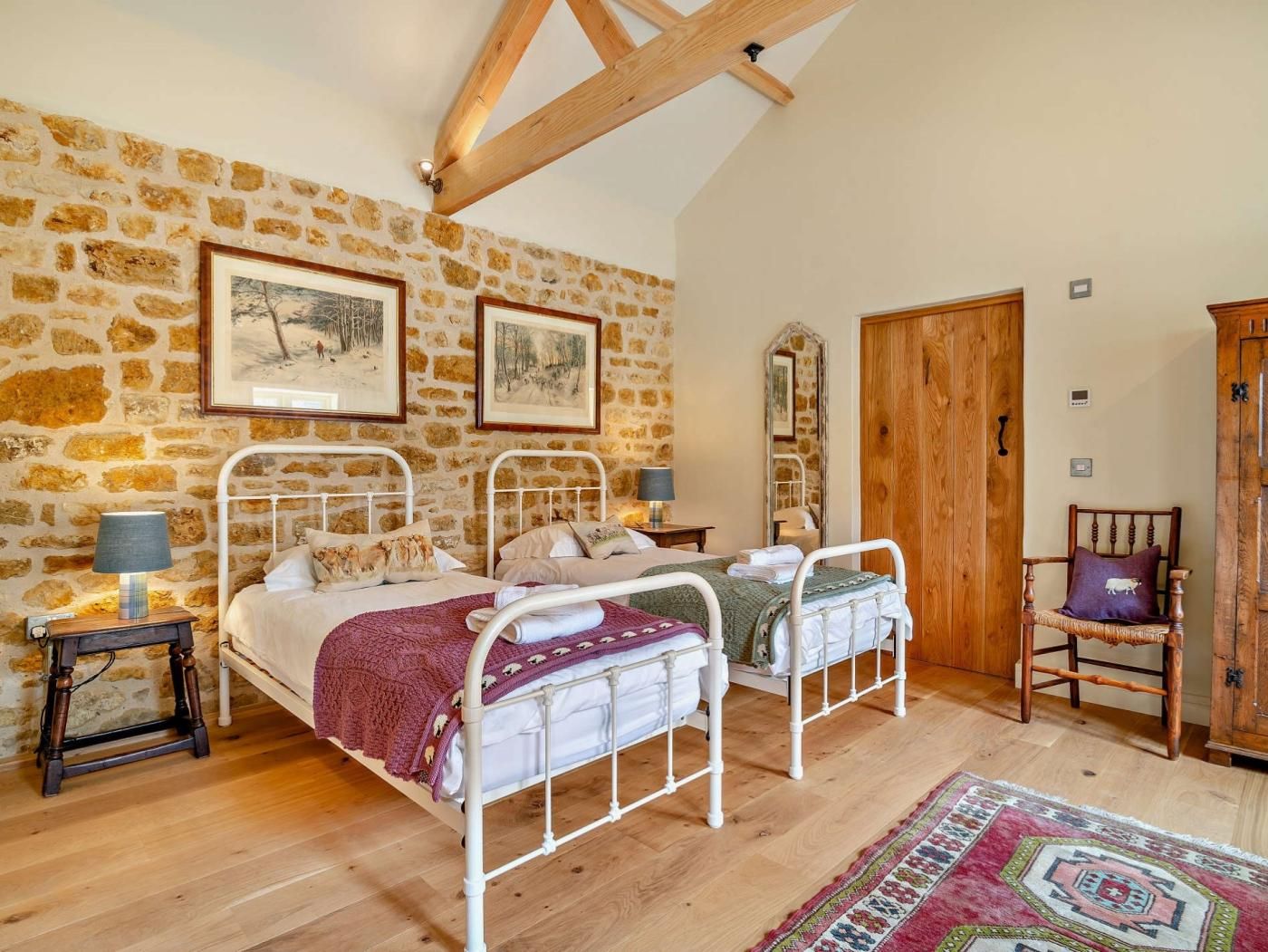 Quarto com duas camas, paredes de pedra, vigas de madeira, quadros, um espelho, um armário de madeira e um tapete estampado.