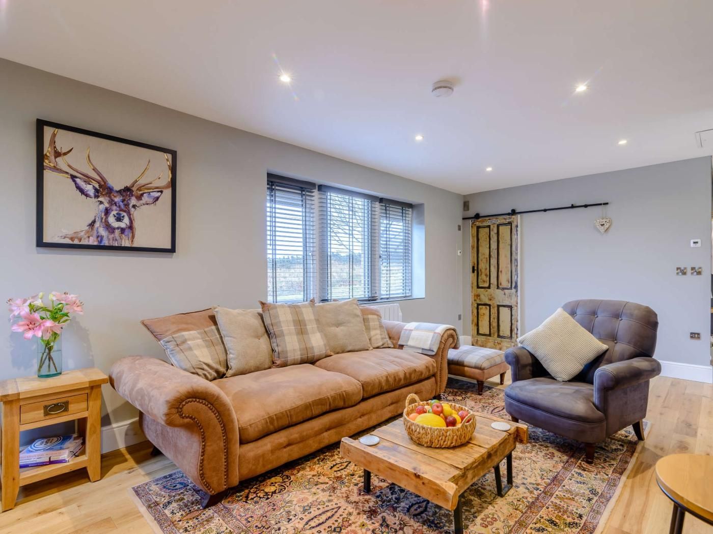 A living room with a brown sofa, a blue armchair, a coffee table with fruit, and a patterned rug.