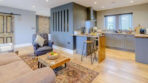 A modern living room with a sofa, a coffee table with fruit, a grey armchair, and a kitchen area with a bar and counter. - 1 Bed in Colne (oc-86513) (Trawden)