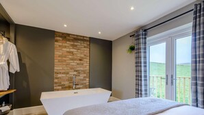 A modern bedroom with a large bed, a freestanding bathtub, and a view of the countryside through a large window. - 1 Bed in Colne (oc-86513) (Trawden)