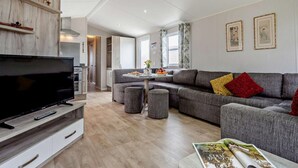 A modern living room with a flat-screen TV, grey sofas, a dining table, and a magazine on a side table. - 3 Bed in Ventnor (oc-94278) (Rookley)
