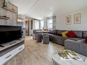 A modern living room with a flat-screen TV, grey sofas, a dining table, and a magazine on a side table. - 3 Bed in Ventnor (oc-94278) (Rookley)