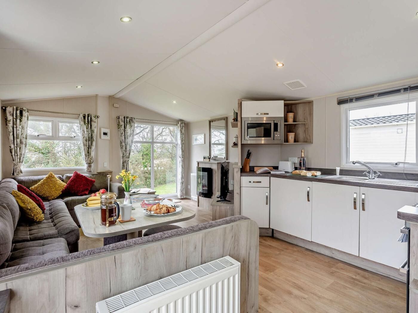 A modern kitchen with white cabinets, a microwave, and a sink. There is a dining area with a table and chairs, and a sofa with cushions.