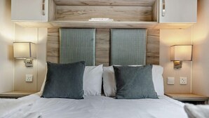 A hotel room with a bed, two bedside tables, wall-mounted lamps, and a wooden headboard. - 3 Bed in Ventnor (oc-94278) (Rookley)