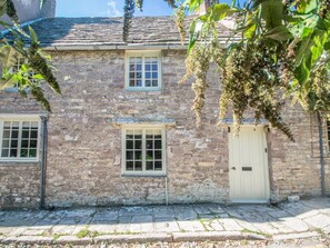 A stone building with two windows and a door, a stone-paved ground, and hanging plants. - 3 Bed in Swanage (oc-dc052) (Worth Matravers)