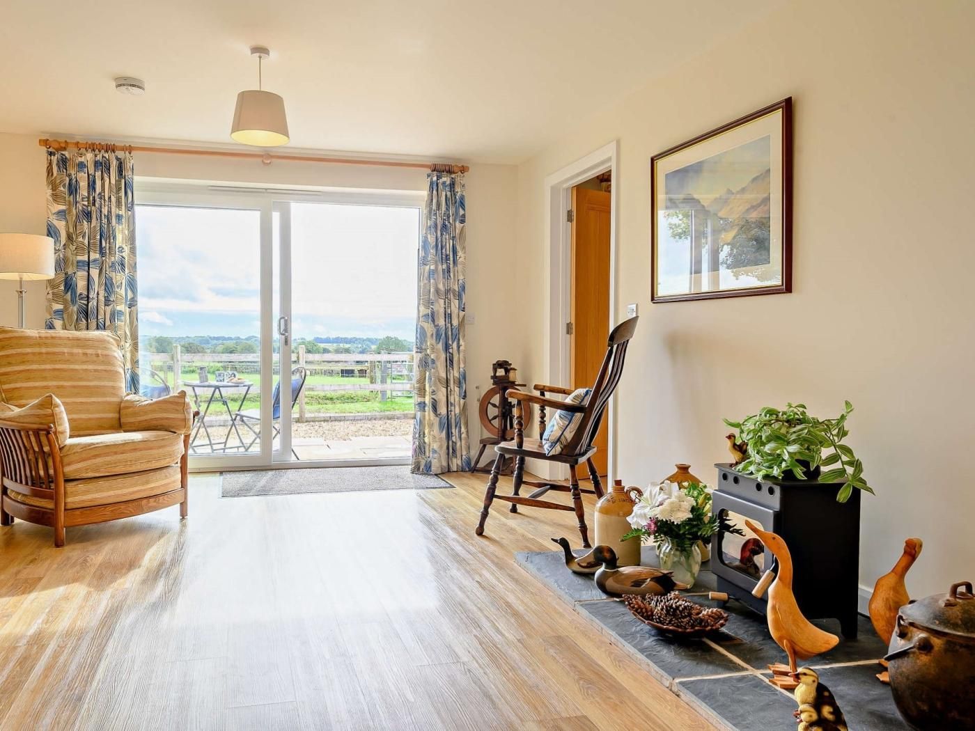 Quarto com piso de madeira, sofá, cadeira, mesa, abajur, pintura e vista através da porta de vidro.