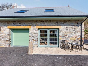 A stone building with a green garage door, a glass door entrance, and a small patio with outdoor furniture. - 1 Bed in Launceston (oc-86675) (South Petherwin)