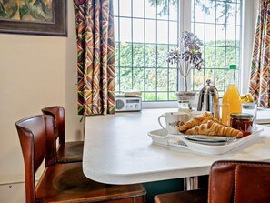 Une table dressée pour le petit-déjeuner, avec un croissant, un jus de fruits et une radio, placée près d’une fenêtre ornée de rideaux.