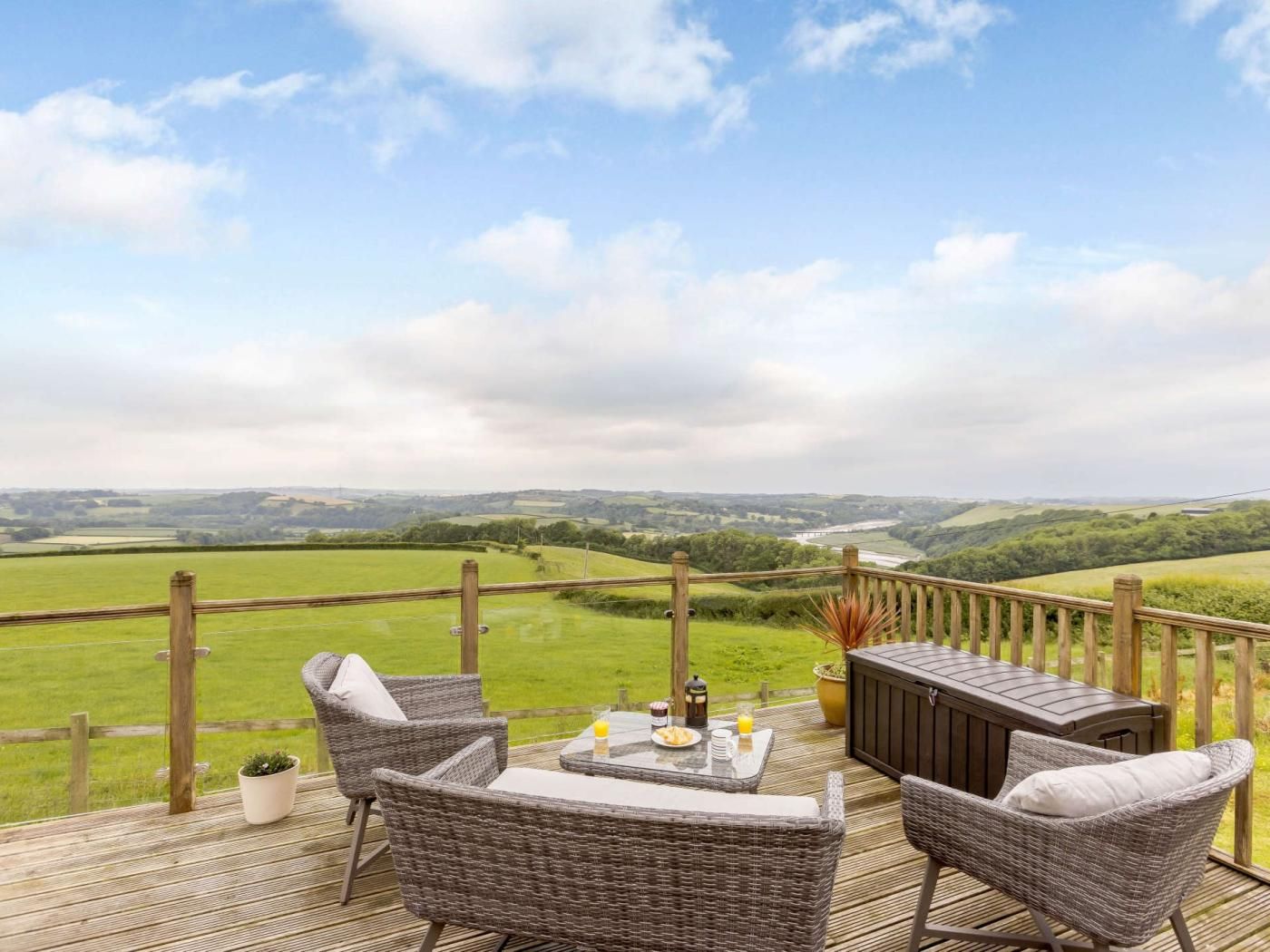 Une terrasse dotée de meubles en osier et d’une table préparée pour le petit-déjeuner, offrant une vue sur un paysage verdoyant et une rivière.
