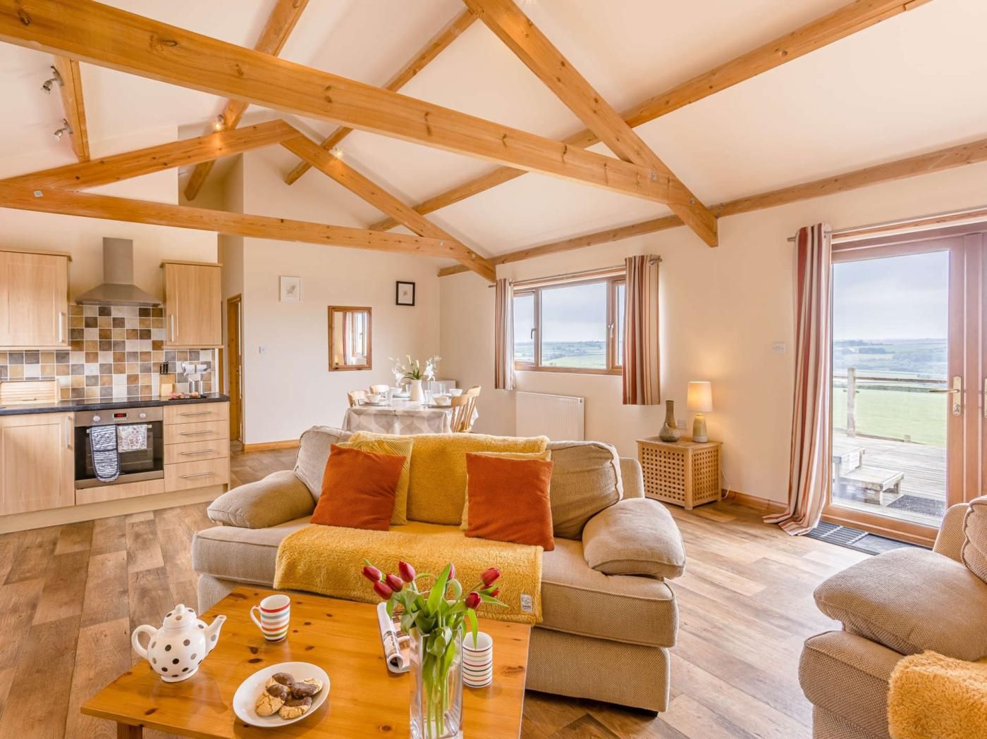 A cozy living room with a sofa, a dining table with a vase of flowers, and a kitchen area with wooden beams.