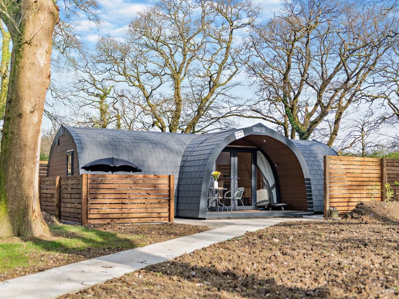 Une structure moderne, semi-ouverte, dotée d’un toit incurvé, d’une clôture en bois et d’un chemin pavé menant à un espace de détente avec une table et des chaises.