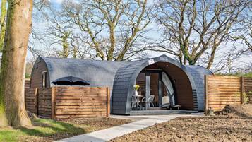 Une structure moderne, semi-ouverte, dotée d’un toit incurvé, d’une clôture en bois et d’un chemin pavé menant à un espace de détente avec une table et des chaises.