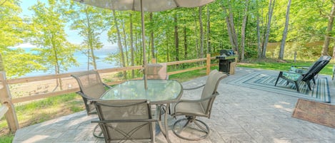 An outdoor patio with a glass table and chairs, a large umbrella, and a view of a lake surrounded by trees.