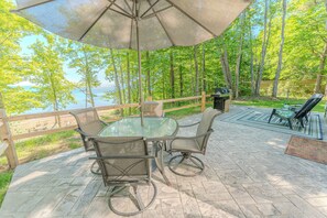 Eine Terrasse im Freien mit Glastisch und -stühlen, einem großen Sonnenschirm und Blick auf einen von Bäumen gesäumten See.